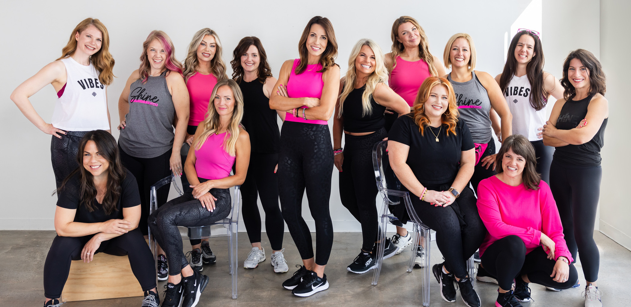 A group of 15 women, some standing and others seated, pose together indoors. They are wearing coordinated activewear in black, pink, and white colors. All are smiling at the camera.