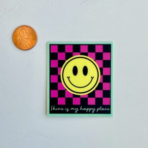 A penny beside a SHiNE & UPLIFT Sticker featuring a yellow smiley face on a pink and black checkerboard background with the text "Shine is my happy place.